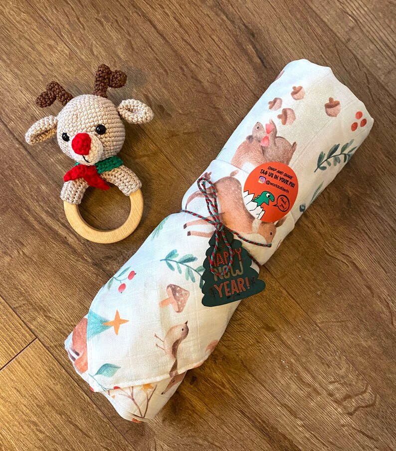 May include: A crocheted reindeer rattle with a wooden ring and a rolled-up white baby blanket featuring woodland animal illustrations. The blanket is tied with twine and a green "Happy New Year!" tag.