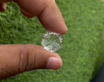 Puede incluir: Una gema de cristal clara, redonda y facetada, sostenida entre los dedos. La gema tiene un patrón detallado en forma de estrella tallado en su superficie. El fondo es verde borroso, lo que sugiere un entorno natural.