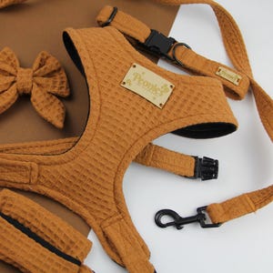 May include: A dog accessory set in a warm brown waffle fabric. The set includes a harness, lead, bow tie, and waste bag dispenser. The harness has a wooden tag with the brand name. The lead and harness have black plastic buckles.
