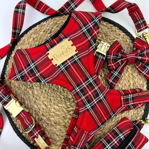 May include: A red plaid dog harness, leash, collar, bow tie, and waste bag set. The set includes gold-colored metal hardware and a wooden tag with the word "Peonies". The items are arranged on a woven mat.