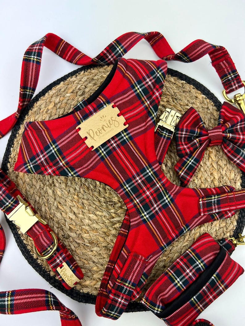 May include: A red plaid dog harness, leash, collar, bow tie, and waste bag set. The set includes gold-colored metal hardware and a wooden tag with the word "Peonies". The items are arranged on a woven mat.