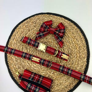 May include: A red and black plaid dog collar, leash, bow tie, and waste bag holder set. The collar and leash have gold-colored hardware. The bow tie is a matching plaid pattern. The waste bag holder is rectangular with a black elastic band.