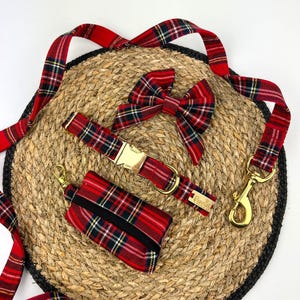 May include: A red plaid dog collar, leash, bow tie, and waste bag dispenser set. The collar has a gold buckle, and the leash has a gold-colored clasp. The set is arranged on a woven mat with a black border.
