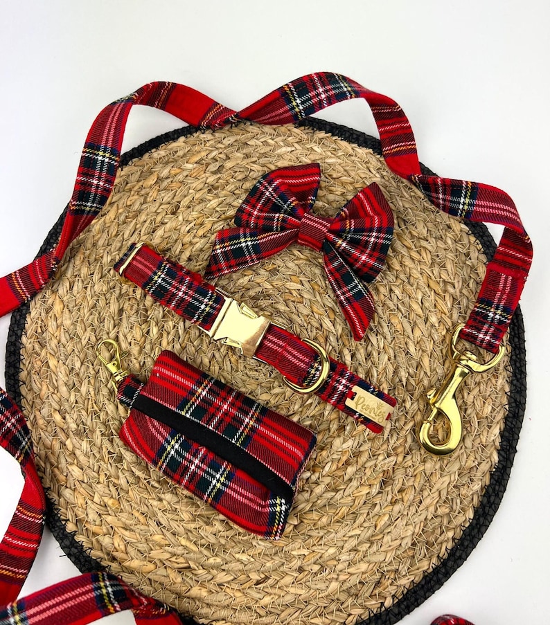 May include: A red plaid dog collar, leash, bow tie, and waste bag dispenser set. The collar has a gold buckle, and the leash has a gold-colored clasp. The set is arranged on a woven mat with a black border.