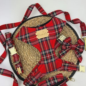 May include: A red plaid dog harness, collar, leash, bow tie, and waste bag set. The set includes gold-colored metal hardware and a wooden tag that reads "Peonies". The items are arranged on a woven mat.
