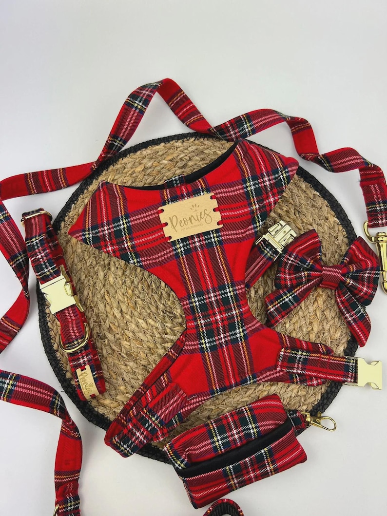 May include: A red plaid dog harness, collar, leash, bow tie, and waste bag set. The set includes gold-colored metal hardware and a wooden tag that reads "Peonies". The items are arranged on a woven mat.