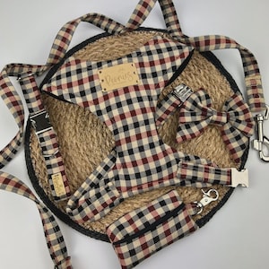 May include: A dog harness set with a matching leash, bow tie, and waste bag dispenser. The set features a black, red, and cream-colored checkered pattern. The harness and leash have metal hardware. The set is displayed on a woven mat.