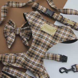 May include: A dog harness, collar, lead, and bow tie set in a brown, black, and white plaid pattern. The harness has a black buckle and a small tag that reads "Peonies". The lead has a black metal clasp.