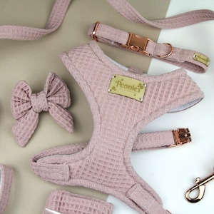 May include: A dog accessory set in a soft pink waffle weave fabric. The set includes a harness, lead, collar, bow, and waste bag holder. The hardware is rose gold. The harness has a tag that says "Peonies".