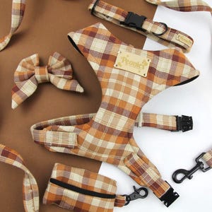 May include: A dog harness set in a plaid pattern of brown, tan, and orange. The set includes a harness, leash, collar, bow tie, and waste bag dispenser. The harness has a wooden tag that reads "Peonies".