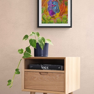 May include: A framed painting depicting two figures walking through a desert landscape with cacti and palm trees. The painting is displayed on a wooden bedside table with a book titled "Vogue" on top.