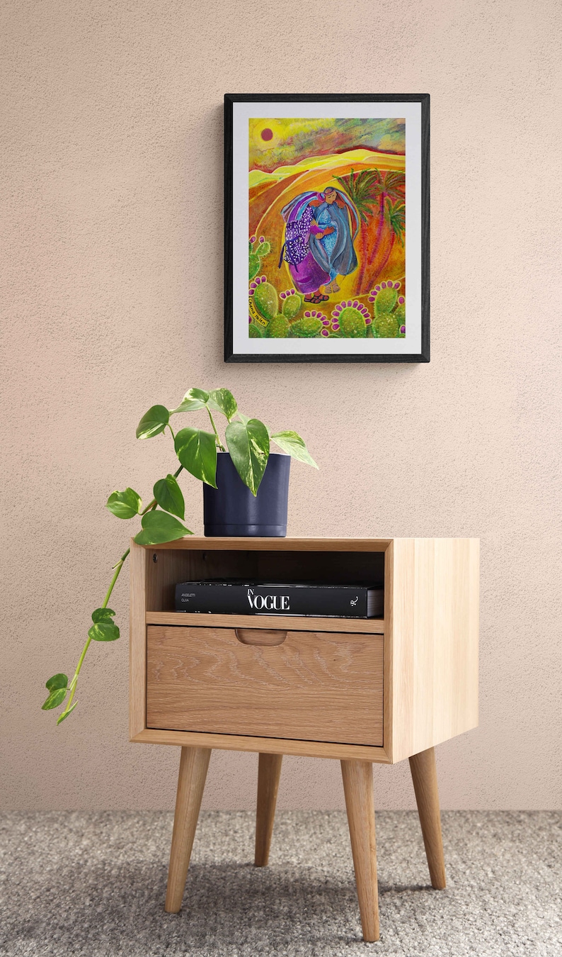May include: A framed painting depicting two figures walking through a desert landscape with cacti and palm trees. The painting is displayed on a wooden bedside table with a book titled "Vogue" on top.