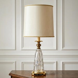 May include: A table lamp featuring a tall, cylindrical cream-coloured fabric shade with gold braided trim. The base is constructed from clear, vertically faceted glass with a tapered shape, supported by a polished gold-tone metal stand and base.