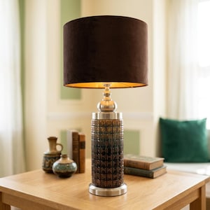 May include: A table lamp featuring a cylindrical base with a dark brown, textured geometric pattern and a metallic silver finish at the top and bottom. The lamp is topped with a solid dark brown velvet drum shade that emits a warm golden glow.