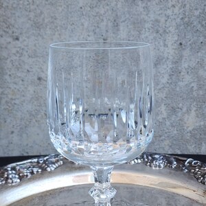 May include: A clear crystal wine glass with vertical and diamond-shaped cut patterns. The glass sits on a decorative silver tray, reflecting the light. The stem has a faceted design. The background is a mottled gray.