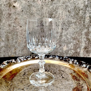 May include: A clear, cut crystal wine glass with a detailed pattern. The glass sits on a silver-colored tray with an ornate design. The glass has a round base, a stem with a decorative element, and a flared bowl.