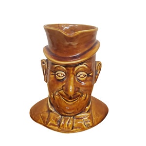 May include: A brown ceramic pitcher shaped like a man's head wearing a brown hat. The man has a mustache and a friendly smile.