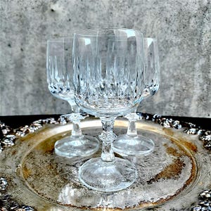 May include: Four clear crystal wine glasses with intricate vertical and diamond-shaped cut patterns. The glasses sit on a silver-colored, ornate tray. The stems have a decorative design. The background is a textured gray.