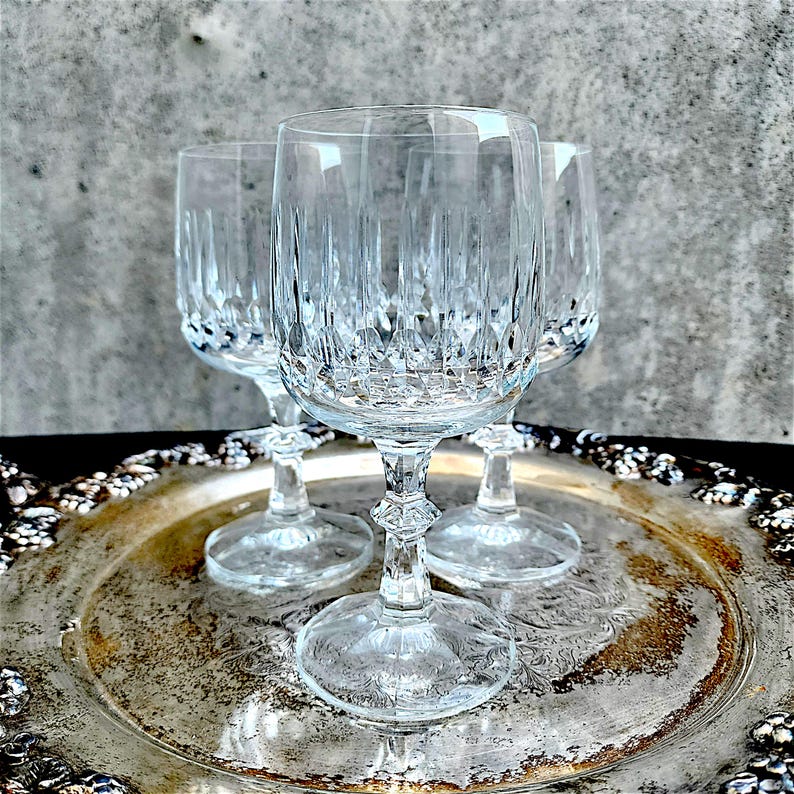 May include: Four clear crystal wine glasses with intricate vertical and diamond-shaped cut patterns. The glasses sit on a silver-colored, ornate tray. The stems have a decorative design. The background is a textured gray.