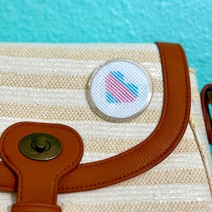 May include: A white striped canvas bag with a brown leather flap and a small, round, clear plastic pin with a pink and blue heart design.
