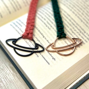 May include: Two book marks with a planet design. One bookmark is black and the other is gold. The bookmarks are attached to a pink and green cord.
