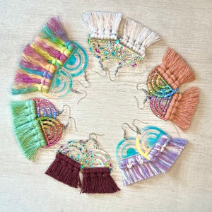 May include: A set of 10 rainbow-themed earrings with colorful tassels. Each earring features a clear acrylic half-circle with a rainbow glitter design and a tassel made of colorful yarn.