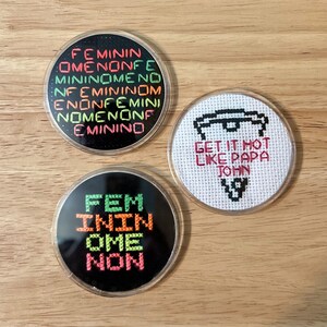 May include: Three round coasters with embroidered designs. The first coaster has a black background with the words "FEMININ OMENONFE MININOMENO NFEMININOM ENONFEMINI NOMENONF EMININO" embroidered in green, orange, and red. The second coaster has a white background with a black and white embroidered design of a pizza slice and the words "GET IT HOT LIKE PAPA JOHN" embroidered in red. The third coaster has a black background with the words "FEM NINI OME NON" embroidered in green, orange, and red.