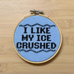 May include: A cross-stitch embroidery hoop with the text "I LIKE MY ICE CRUSHED" in black on a light blue background. The embroidery is framed by a wooden hoop and set against a wooden surface.