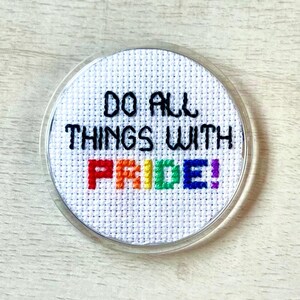 May include: A round, cross-stitched design with the text "DO ALL THINGS WITH PRIDE!" in black thread. The word "PRIDE" is stitched in rainbow colors. The design is encased in a clear, circular frame, suitable for a pin or magnet.