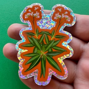 May include: A holographic sticker featuring a stylized illustration of a plant with white flowers and green leaves on an orange background.