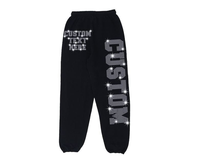 K&ouml;nnte beinhalten: Schwarze Jogginghose mit einem Strass-Design am rechten Bein, das "CUSTOM" sagt. Das linke Bein hat den Text "CUSTOM TEXT HERE" in Strasssteinen.