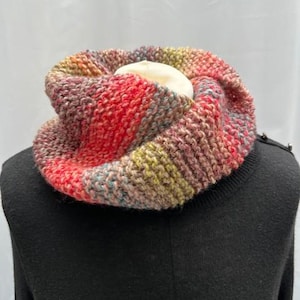 May include: A hand-knitted cowl in a mix of colors including red, pink, green, and blue. The cowl is made of thick yarn and has a textured appearance. The item is displayed on a black surface.