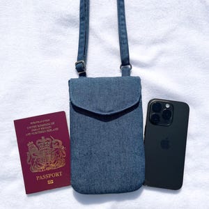 May include: A blue denim crossbody phone bag with a flap and adjustable strap. A black smartphone and a burgundy passport are next to the bag. The passport has gold lettering and the words "Passport" and "European Union" printed on it.