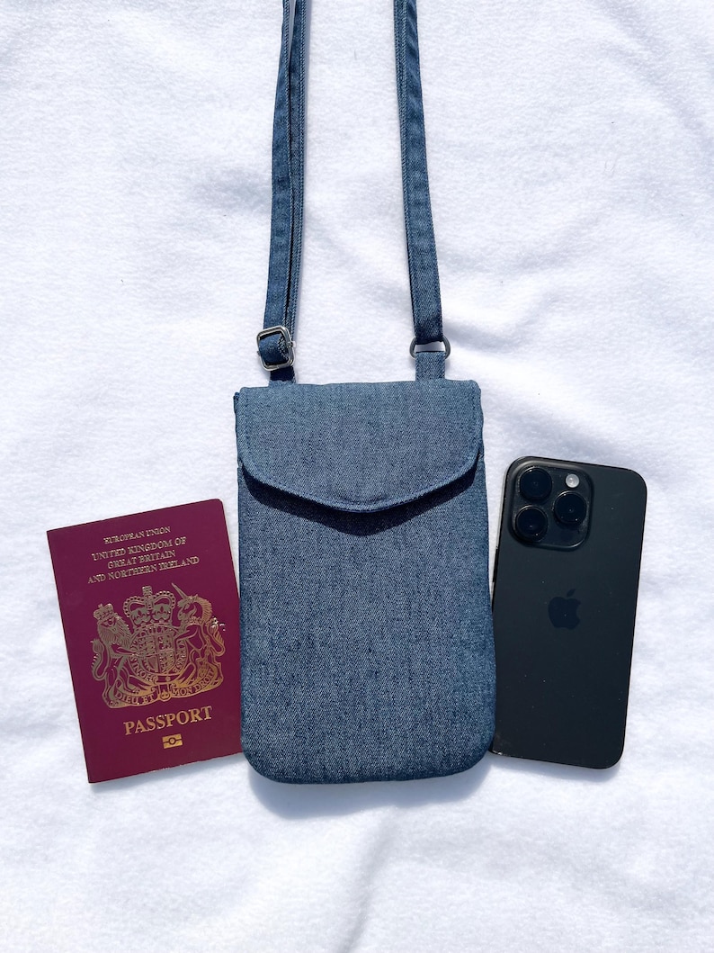 May include: A blue denim crossbody phone bag with a flap and adjustable strap. A black smartphone and a burgundy passport are next to the bag. The passport has gold lettering and the words "Passport" and "European Union" printed on it.