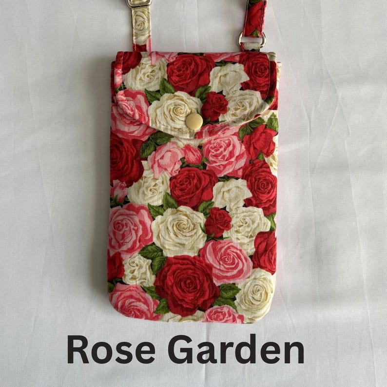 Puede incluir: Bolso para tel&eacute;fono con estampado floral con un patr&oacute;n de rosas rojas, rosas y blancas. El bolso tiene un cierre de solapa con un bot&oacute;n y una correa ajustable. El texto "Rose Garden" est&aacute; impreso en la parte inferior.