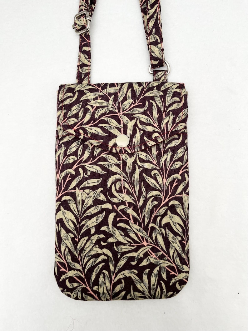 May include: A small burgundy fabric phone pouch with a leafy pattern and a white button closure. The pouch has a long adjustable strap.