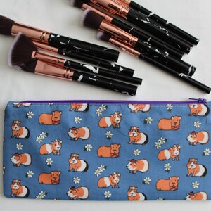 Puede incluir: Una bolsa de maquillaje azul con cremallera con un patr&oacute;n de conejillos de indias con sombreros y pajaritas. Los conejillos de indias son naranja y blanco y el fondo es azul claro con flores blancas.