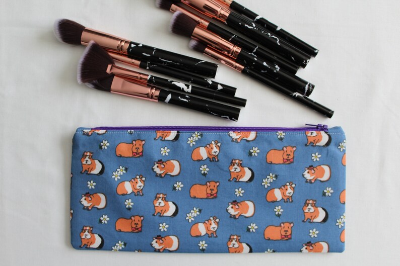 Puede incluir: Una bolsa de maquillaje azul con cremallera con un patr&oacute;n de conejillos de indias con sombreros y pajaritas. Los conejillos de indias son naranja y blanco y el fondo es azul claro con flores blancas.