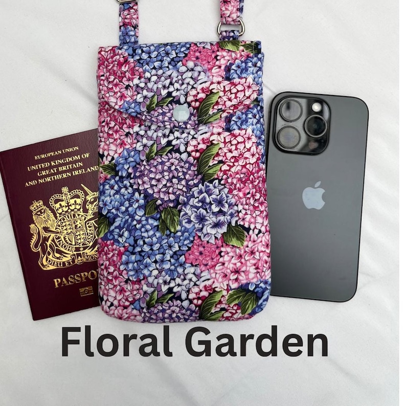 May include: A floral print phone crossbody bag with a matching strap. The bag features a button closure and a design of pink, purple, and blue flowers. A black smartphone and a passport are next to the bag. The text "Floral Garden" is at the bottom.