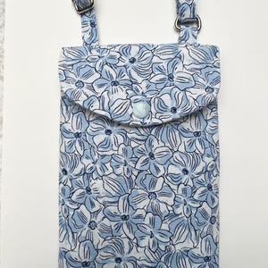 Puede incluir: Un bolso cruzado para tel&eacute;fono con estampado floral azul claro con una correa ajustable a juego. El bolso tiene un cierre de solapa con un bot&oacute;n y herrajes plateados. El estampado floral est&aacute; compuesto por flores azules sobre un fondo blanco.