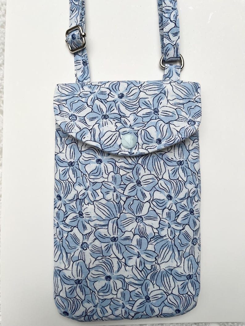 Puede incluir: Un bolso cruzado para tel&eacute;fono con estampado floral azul claro con una correa ajustable a juego. El bolso tiene un cierre de solapa con un bot&oacute;n y herrajes plateados. El estampado floral est&aacute; compuesto por flores azules sobre un fondo blanco.