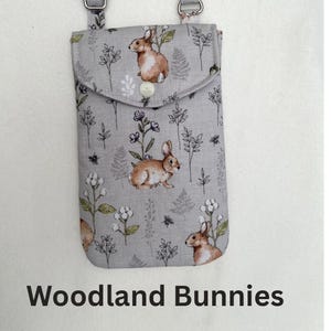 Puede incluir: Una funda de tel&eacute;fono de tela gris con cierre de solapa, con un patr&oacute;n repetido de conejos marrones y elementos florales. El texto "Woodland Bunnies" est&aacute; impreso en la parte inferior. La funda tiene un accesorio de correa plateado.