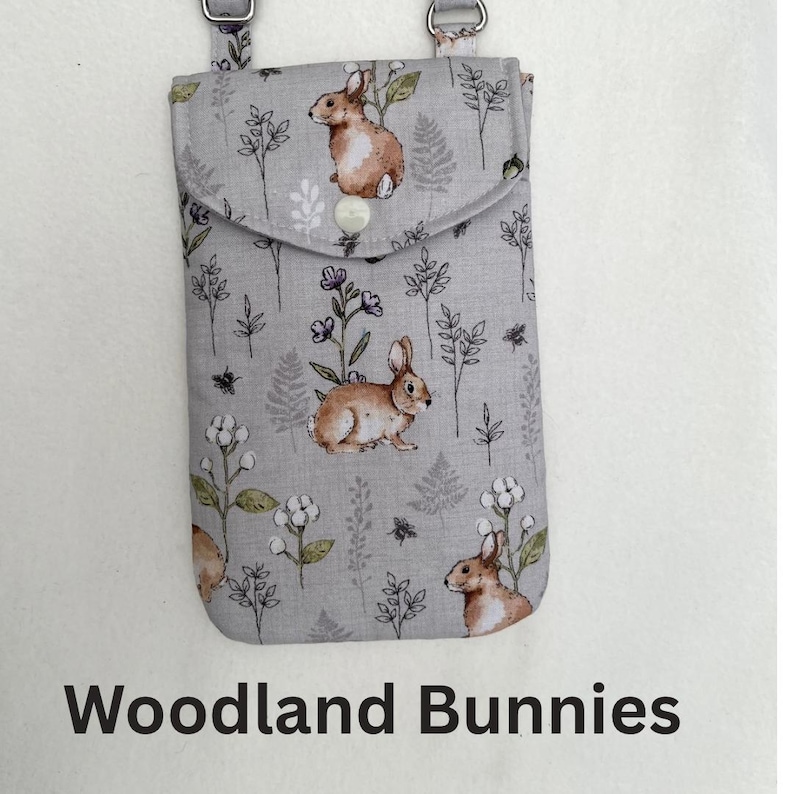 Puede incluir: Una funda de tel&eacute;fono de tela gris con cierre de solapa, con un patr&oacute;n repetido de conejos marrones y elementos florales. El texto "Woodland Bunnies" est&aacute; impreso en la parte inferior. La funda tiene un accesorio de correa plateado.