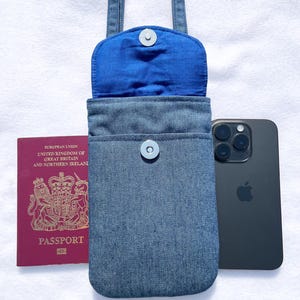 May include: A denim phone bag with a blue interior and a snap closure. A passport and a black smartphone are next to the bag. The bag has a strap for carrying over the shoulder.