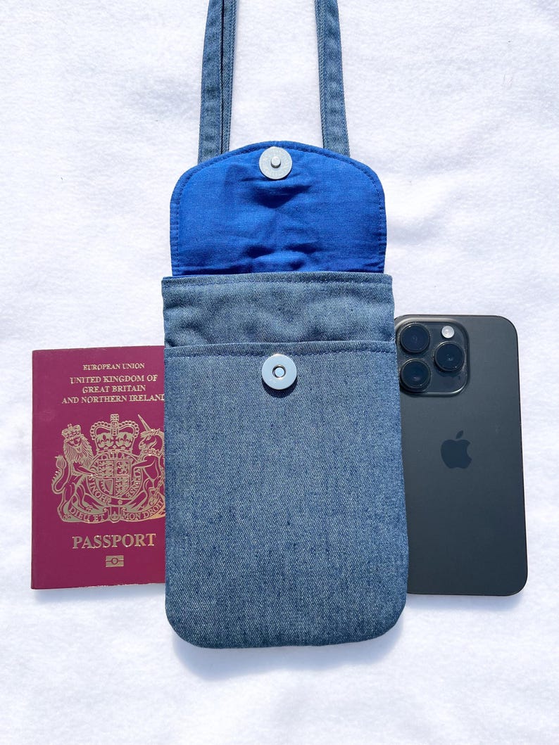 May include: A denim phone bag with a blue interior and a snap closure. A passport and a black smartphone are next to the bag. The bag has a strap for carrying over the shoulder.