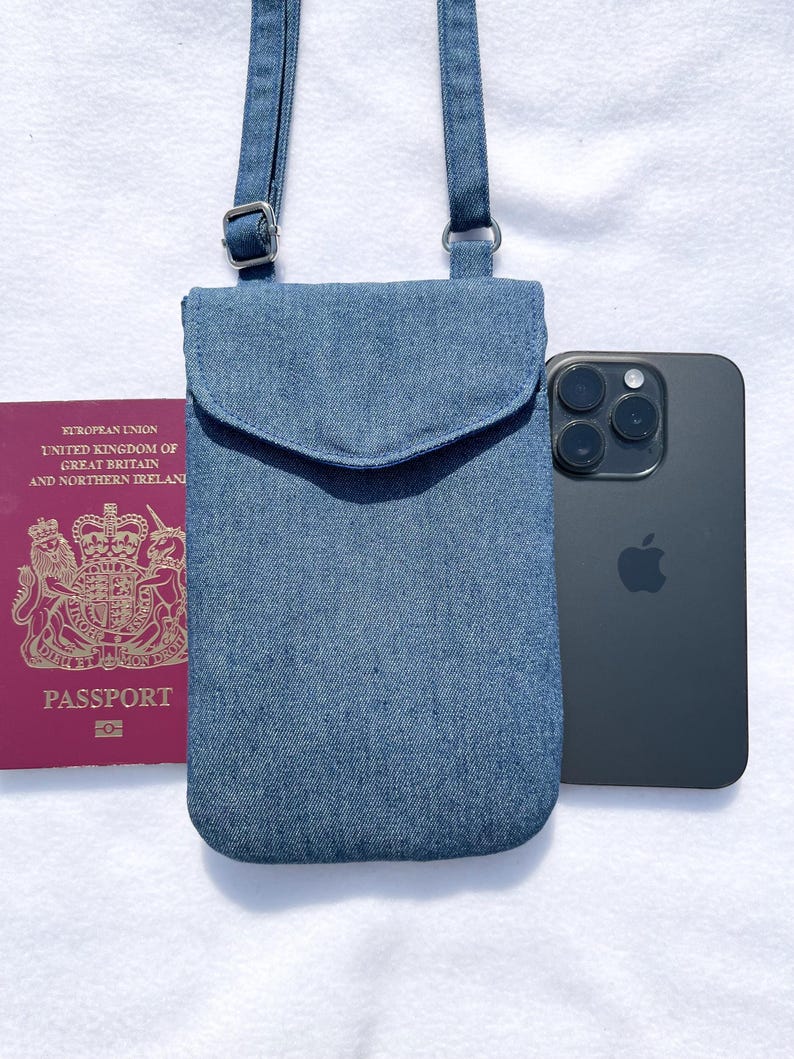 May include: A blue denim crossbody phone bag with a flap and adjustable strap. A black smartphone and a UK passport are next to the bag. The bag is made of denim fabric.