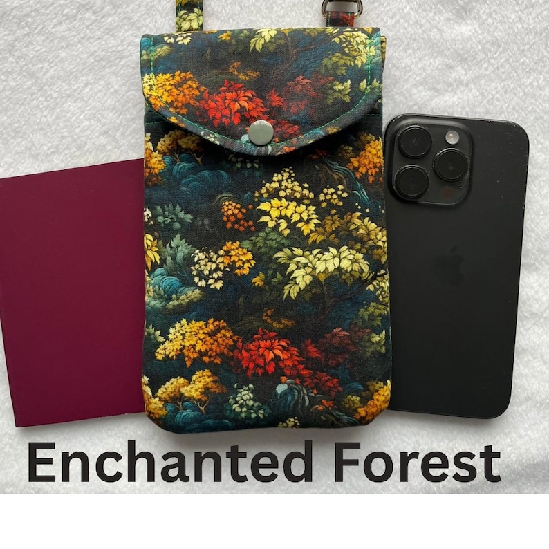 May include: A floral print phone crossbody bag with a flap closure and a button. The bag features a colorful design with red, yellow, and green foliage. A black smartphone and a burgundy item are next to the bag. The text "Enchanted Forest" is at the bottom.