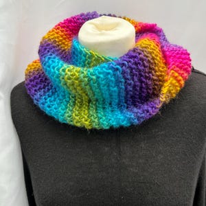 May include: A chunky knit infinity scarf featuring a vibrant rainbow gradient of purple, blue, green, yellow, orange, and pink yarn. The circular scarf has a textured garter stitch pattern and is displayed on a black garment form.