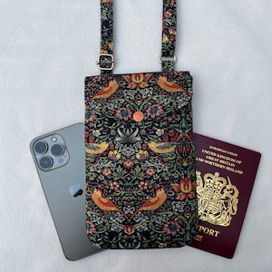 May include: A black phone case with a floral pattern and birds. The case has a long adjustable strap and a flap that closes with a button. The case is lying on a white surface next to a black smartphone and a red passport.
