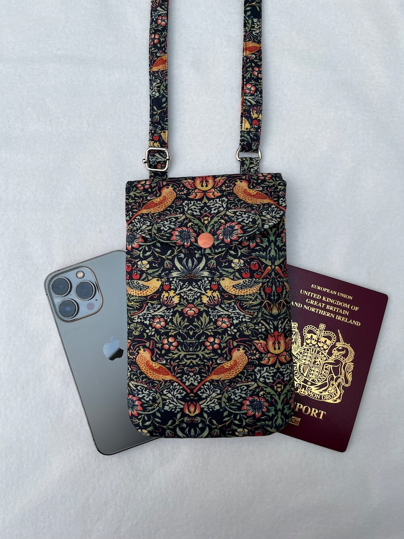 May include: A black phone case with a floral pattern and birds. The case has a long adjustable strap and a flap that closes with a button. The case is lying on a white surface next to a black smartphone and a red passport.
